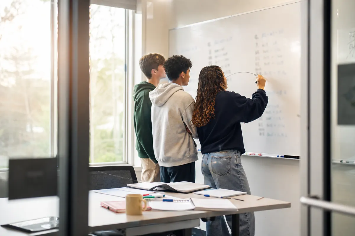 Three friends compare two side-by-side algebra methods on a whiteboard in a glass-walled study room.