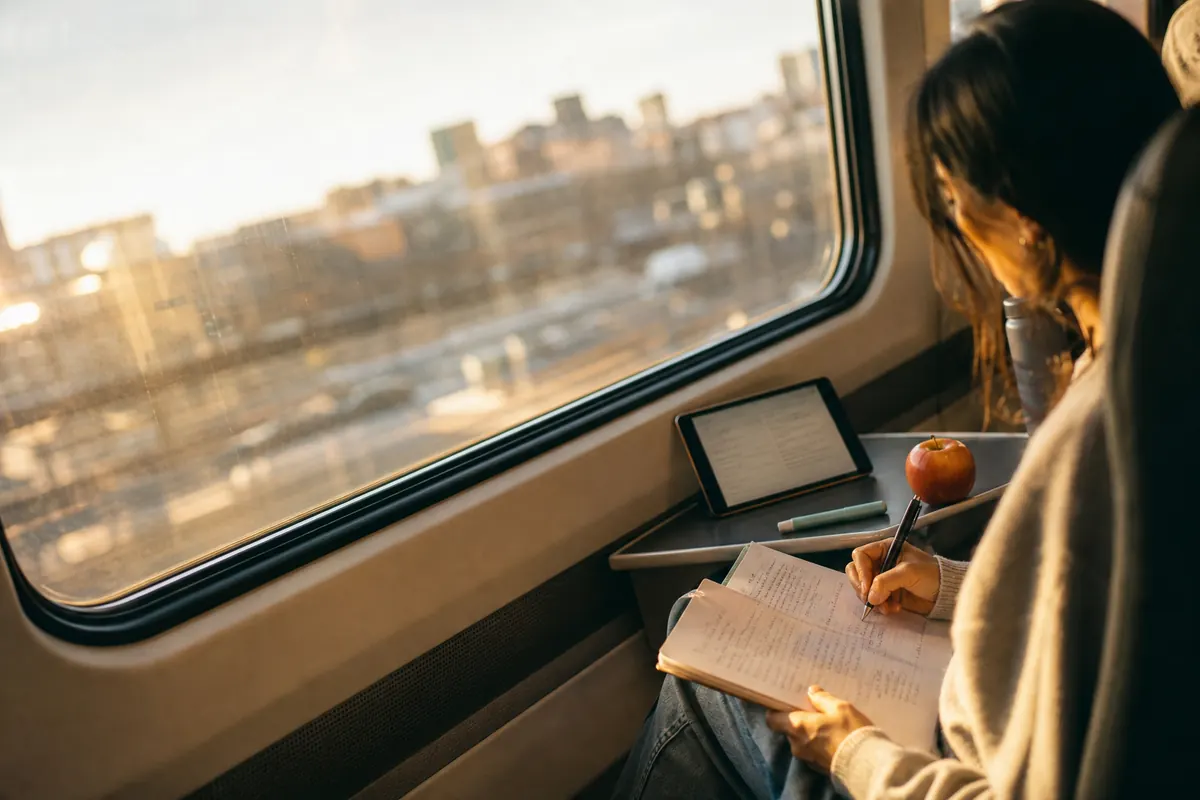 A university student on a train pairs a tablet question bank with answer-free work in a notebook.