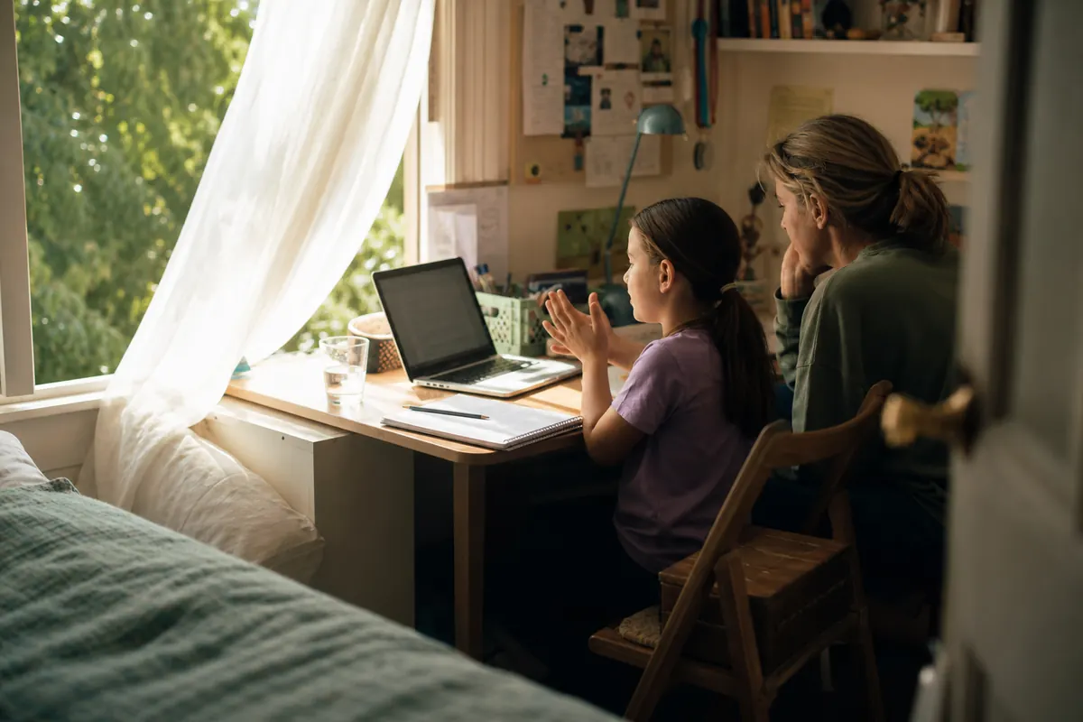 A child and parent at a bedroom desk pause a homework video and predict the next step beside an open notebook.